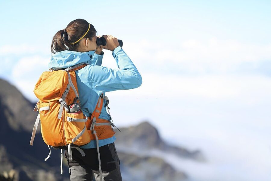 Un excursionist care poartă o jachetă softshell dama se uită cu binoclu și se bucură de o priveliște spectaculoasă de pe vârful muntelui deasupra norilor.