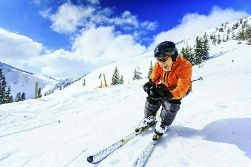 Schior în echipament de turație, în acțiune pe pârtie cu cer albastru și copaci de munte în fundal.