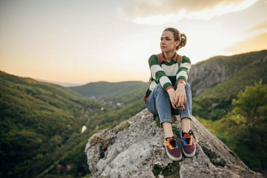 Imagine cu o femeie excursionist așezată pe o stâncă înaltă privind apusul, purtând bocanci de drumeție pentru femei.