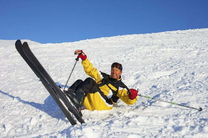 Schior căzut pe zăpadă, râzând și ridicând schiurile de tură în aer pe o pantă.