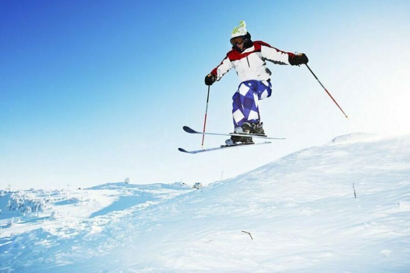 Schior efectuând un salt cu schiuri de tură pe un fundal de cer albastru și teren însorit cu zăpadă.