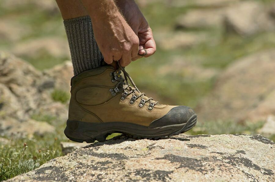 Drumeț legându-și șireturile la bocanci de trekking robusti, purtând șosete de drumeție din lână gri, pregătit pentru aventura montană.