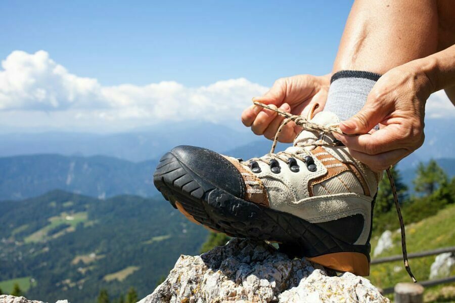 Drumeț legându-și șireturile la bocancii robusti, cu șosete de drumeție gri, pe un vârf de munte cu priveliște panoramică.