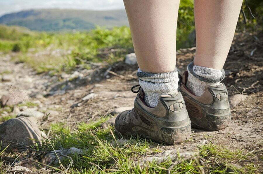 Picioarele unui drumeț în șosete de lână groase și bocanci murdari pe un traseu de munte, cu dealuri verzi în fundal.