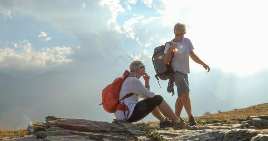 Două femei în pantaloni de drumeție se odihnesc pe un munte, cu soarele strălucind prin norii de deasupra și priveliștea văii de dedesubt.