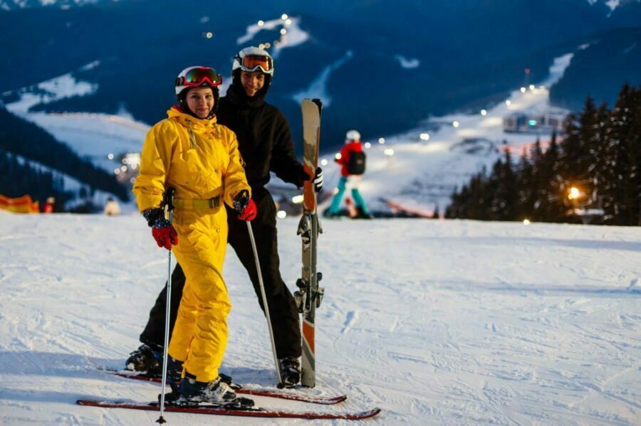 Cuplu de schiori stând pe pârtie seara, cu legături de schiuri și peisaj montan iluminat în fundal