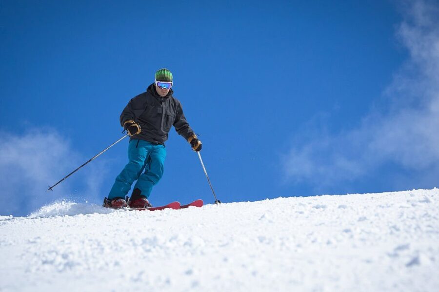 Schior bărbat cu pantaloni de ski turcoaz și jachetă neagră pe cer albastru