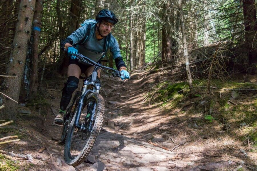 Biciclist de munte în coborâre prin pădure, pe o potecă cu rădăcini în regiunea Dolomiților, Italia.