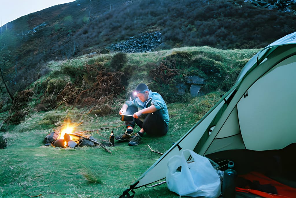 Bărbat folosind o lanternă frontală în timp ce pregătește mâncarea lângă un foc de tabără și cort, la amurg în natură.