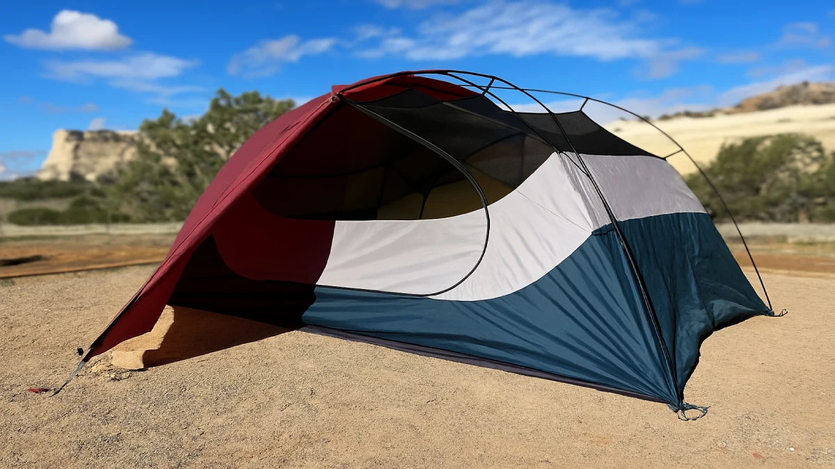 Cort de camping în culori burgundy, gri și albastru închis, montat într-un peisaj arid cu cer senin.