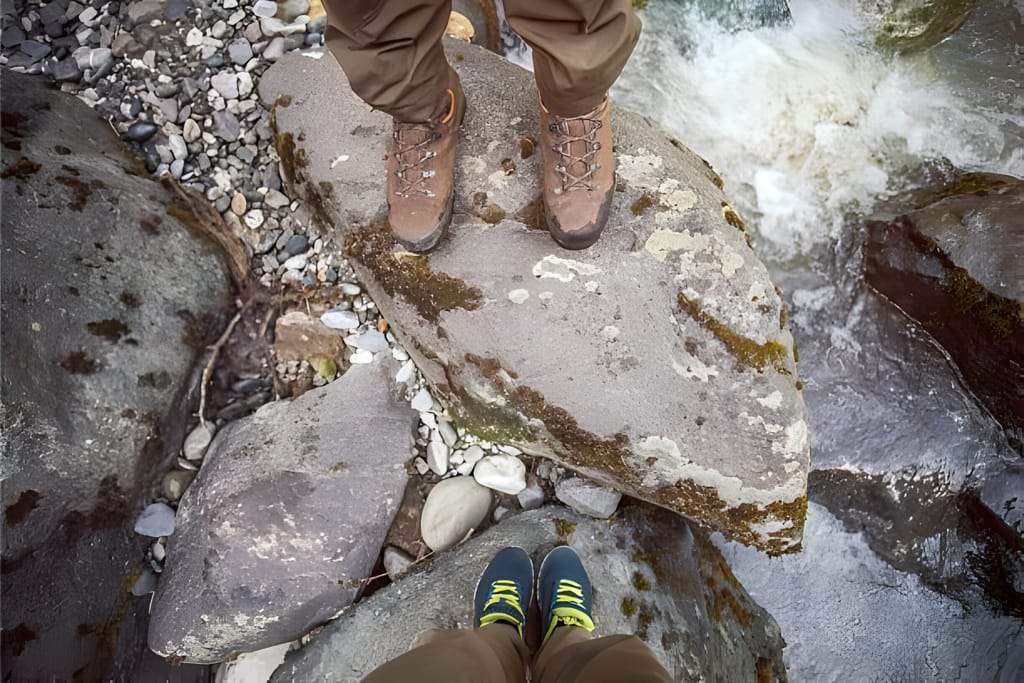 Perspectivă din prima persoană a unor drumeți care traversează un râu pe pietre, cu bocanci de drumeție.