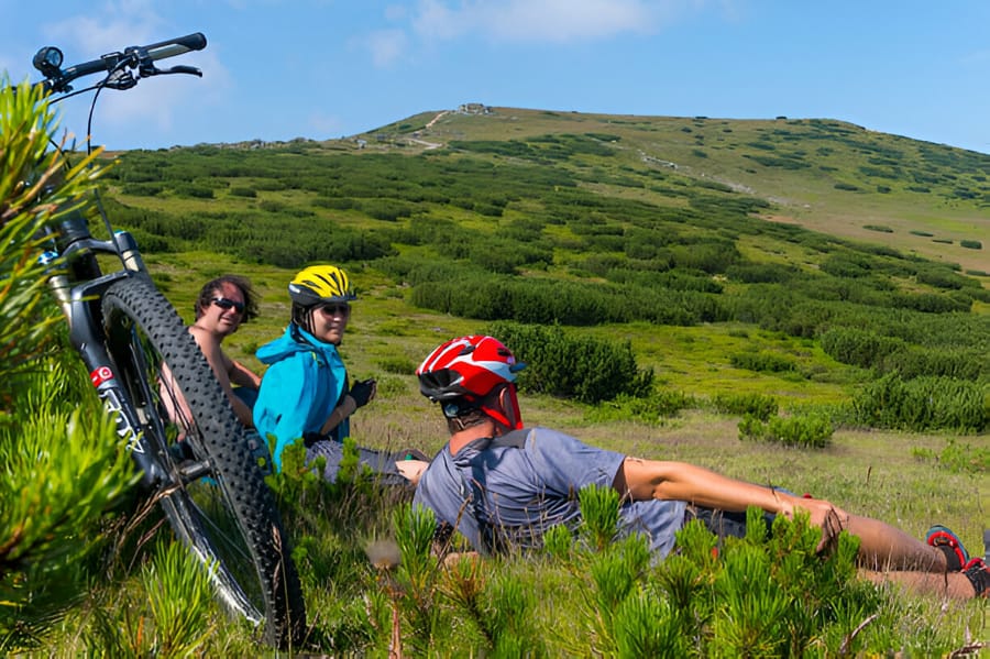 Trei cicliști de mountain bike purtând căști se odihnesc pe un deal verde.
