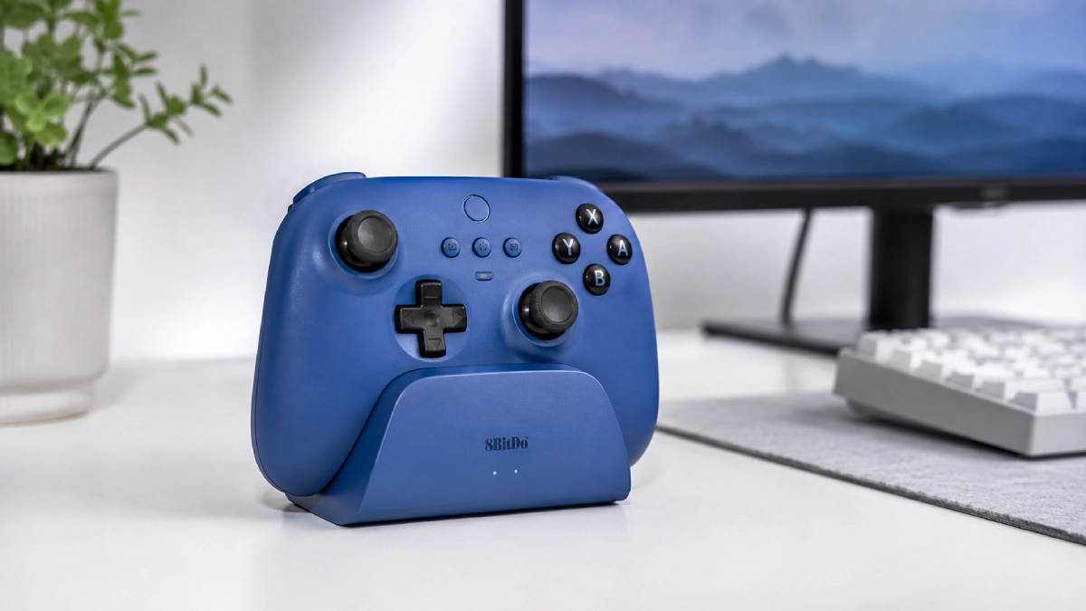 Gamepad 8BitDo Ultimate albastru așezat în dock-ul de încărcare pe un birou deschis, cu monitor estompat în fundal