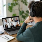 Persoană într-un birou de acasă, purtând căști wireless over-ear în timpul unei videoconferințe pe laptop, cu telefonul alături