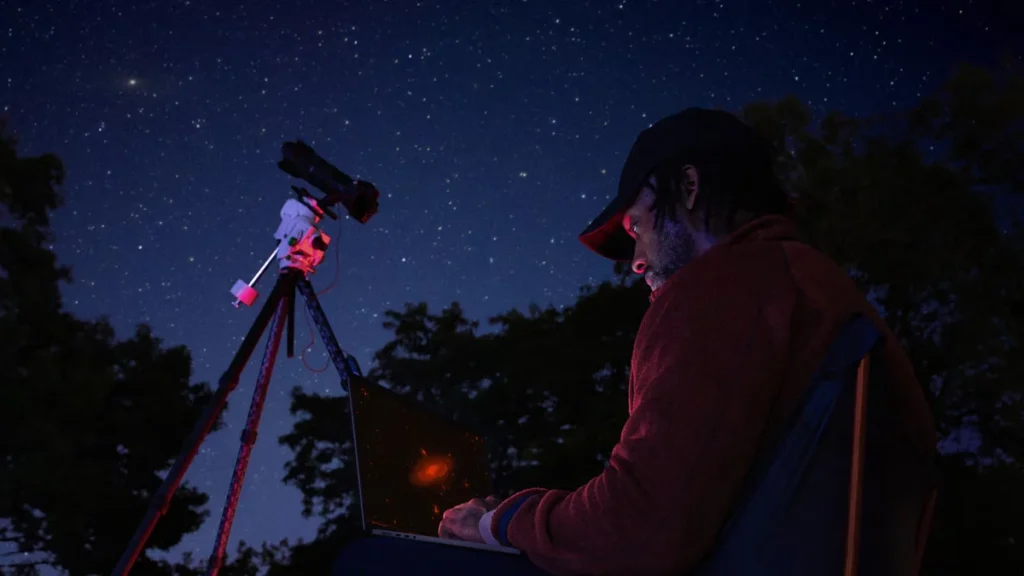 Un bărbat lucrează noaptea pe un MacBook Pro de 14 inch, lângă un telescop montat pe trepied, sub cerul înstelat