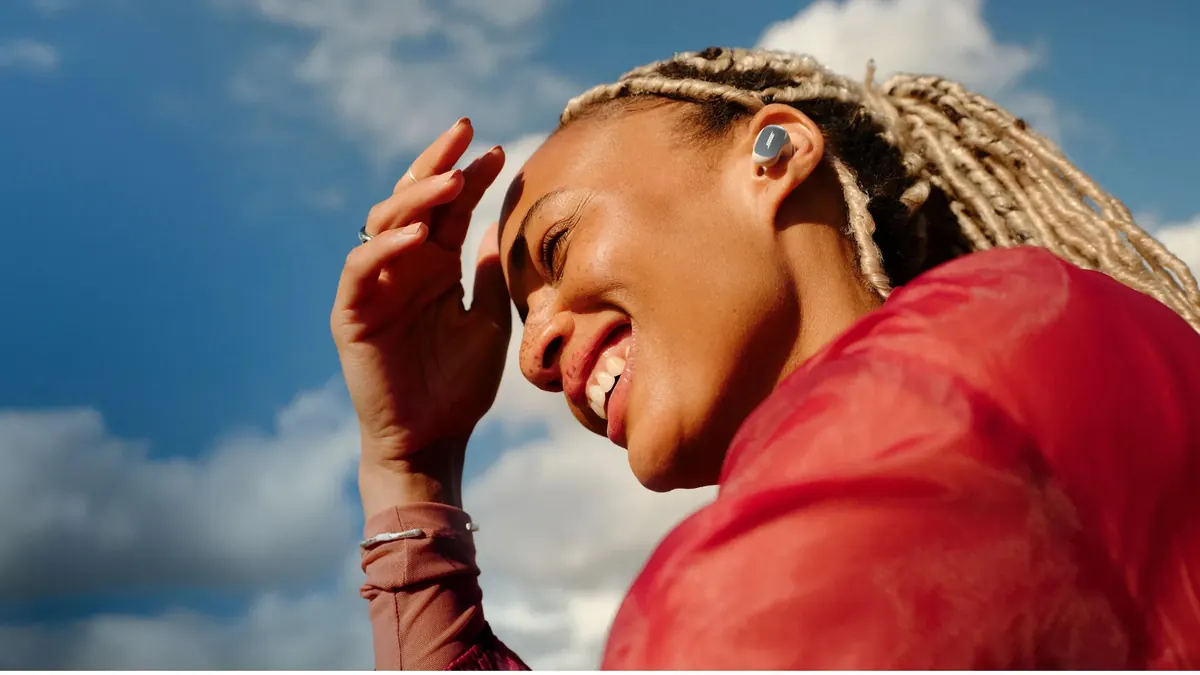 Femeie care zâmbește, cu o mână la tâmplă, purtând o cască in-ear Bose QuietComfort Ultra Earbuds (2nd Gen) în aer liber, pe fundal de cer cu nori