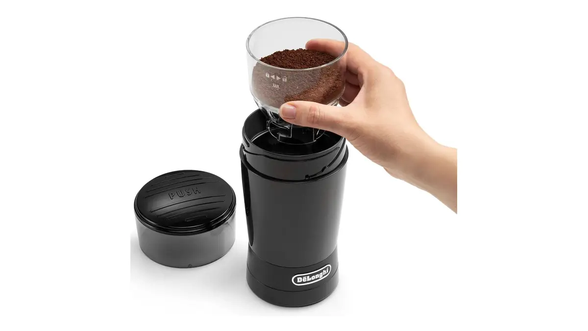 O mână scoate recipientul transparent cu cafea măcinată din râșnița De'Longhi KG200, neagră, cu capacul alături