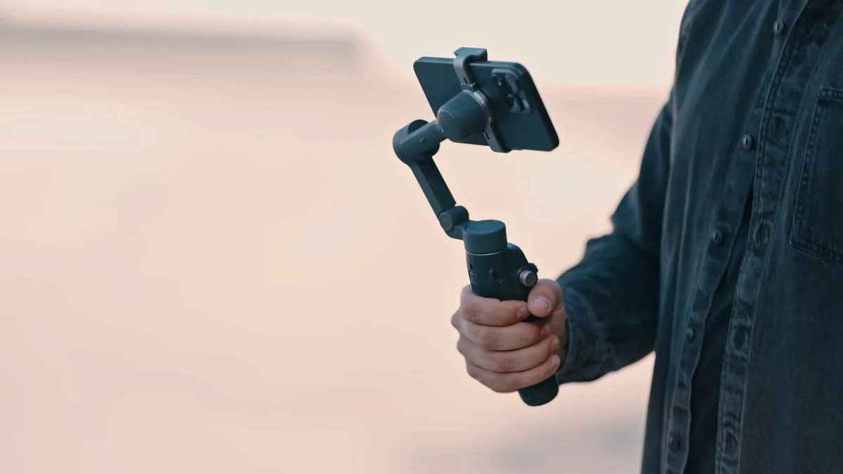 Mână care ține gimbalul DJI Osmo Mobile 8, gri închis, cu un telefon prins în clemă, pe un fundal luminos