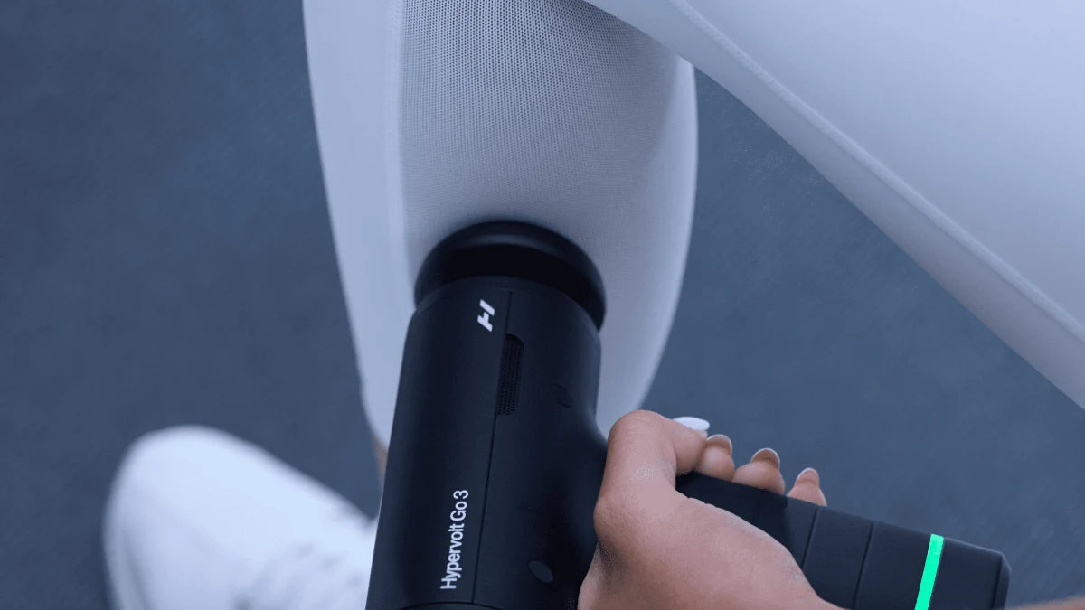 Pistolul de masaj Hyperice Hypervolt Go 3, negru, ținut în mână pe gamba unui sportiv în pantofi albi