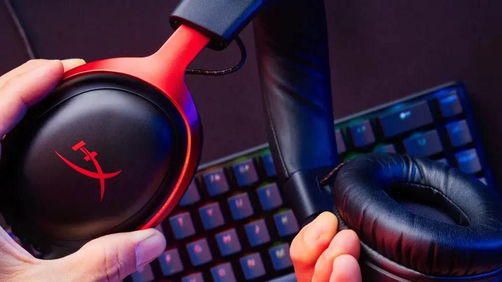 Căștile HyperX Cloud III negru cu roșu, ținute în mână deasupra unei tastaturi de gaming iluminate