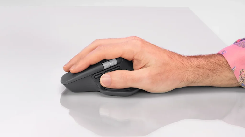 Mână dreaptă care ține mouse-ul Logitech MX Master 4 grafit pe un birou alb, cu degetul mare pe rotița laterală