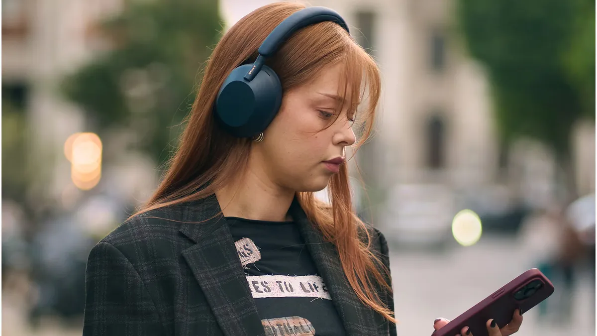 Femeie pe stradă, cu căștile albastre Sony WH-1000XM5 pe cap, uitându-se la telefonul din mână
