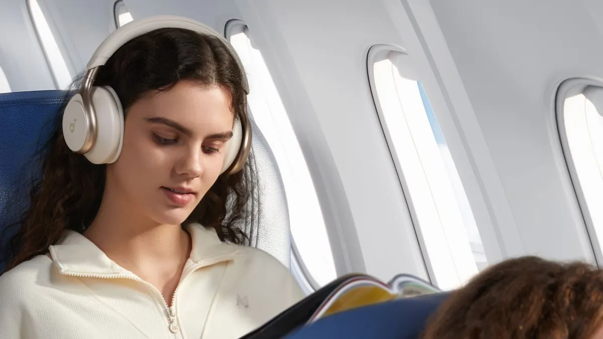 Femeie cu căști Anker Soundcore Space One albe, care citește o revistă pe scaunul albastru al unui avion, lângă geamuri