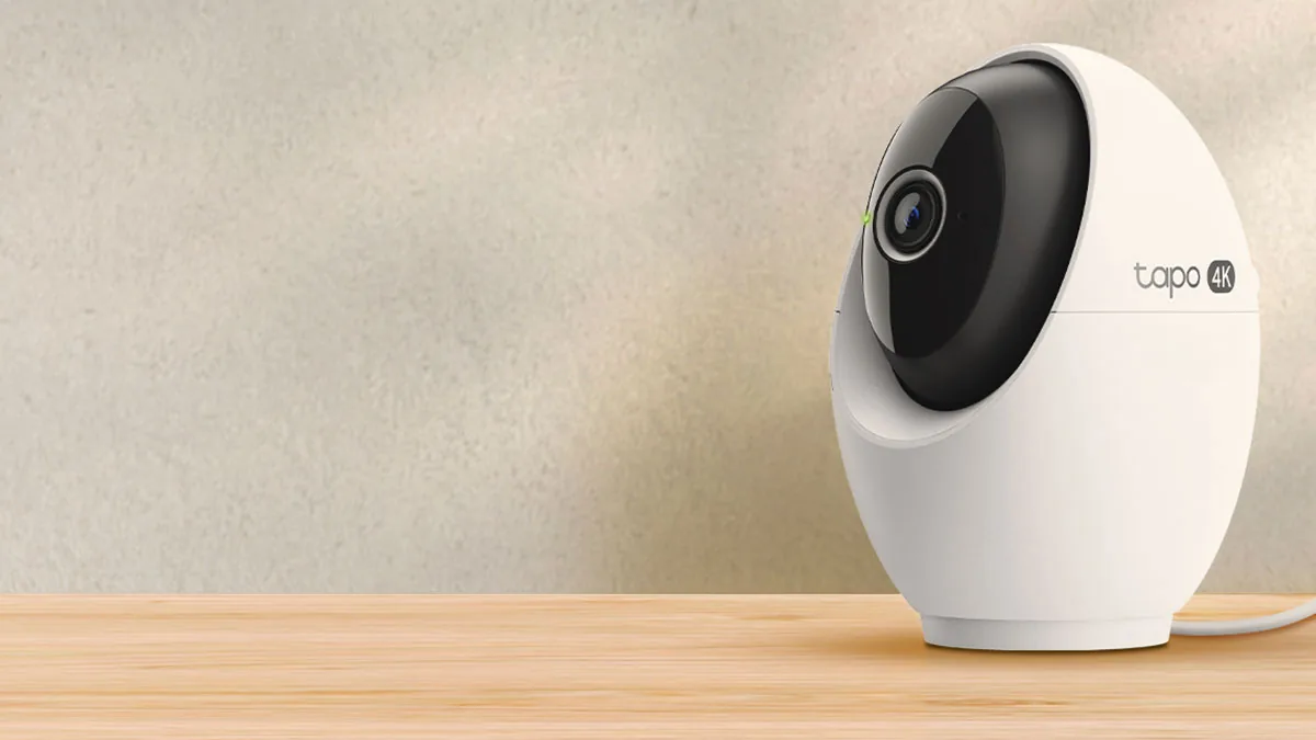 Camera Tapo C260 albă, de aproape, pe un raft din lemn lângă un perete bej, cu obiectivul negru și cablul de alimentare care iese din bază