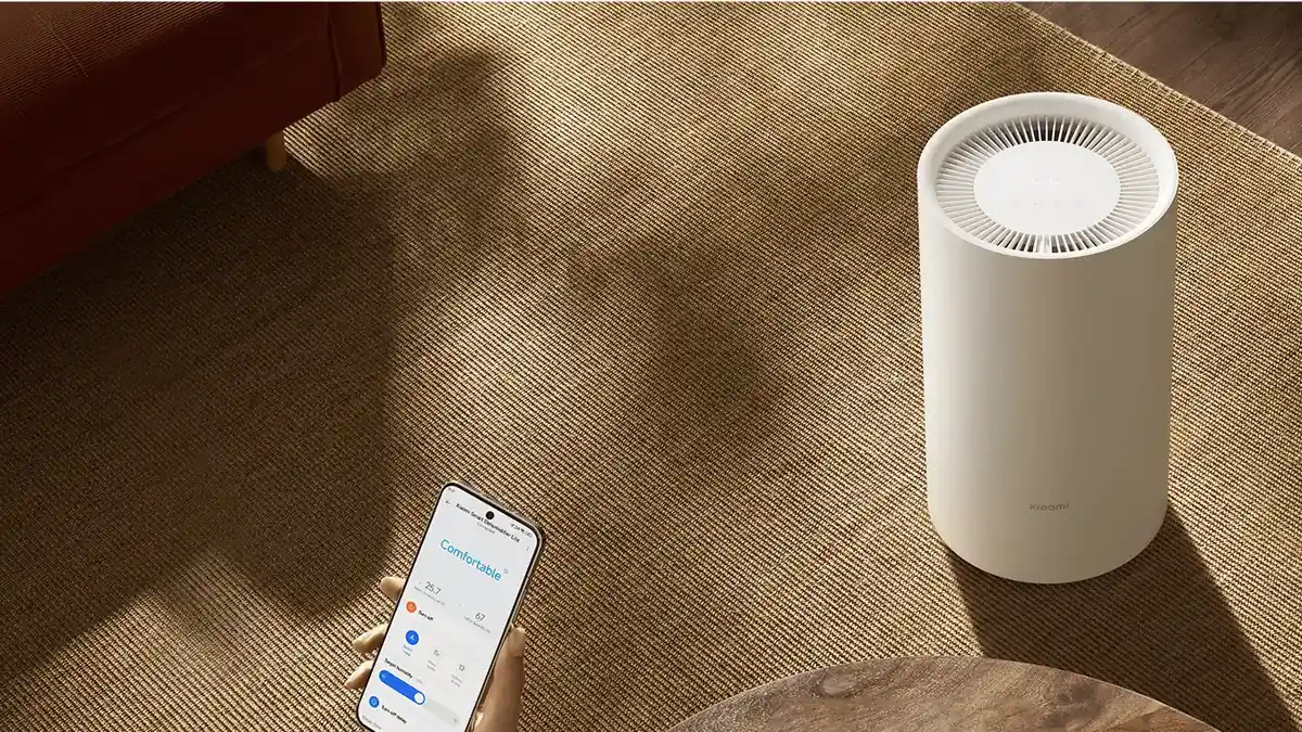 O mână ține un telefon cu aplicația Xiaomi Home deschisă, lângă dezumidificatorul Xiaomi Smart Dehumidifier Lite de pe un covor